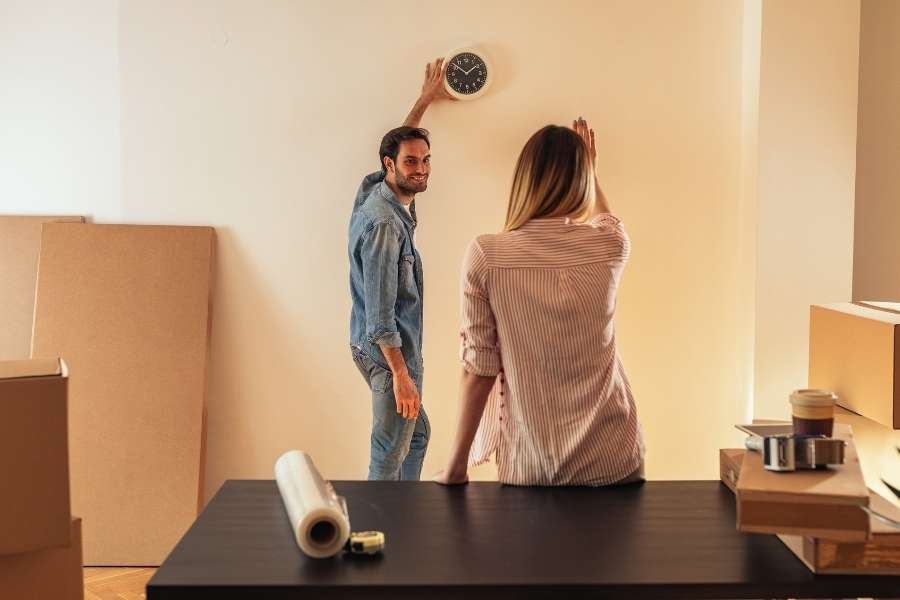 A smiling couple in an empty room surrounded by cardboard boxes, hanging a wall clock.